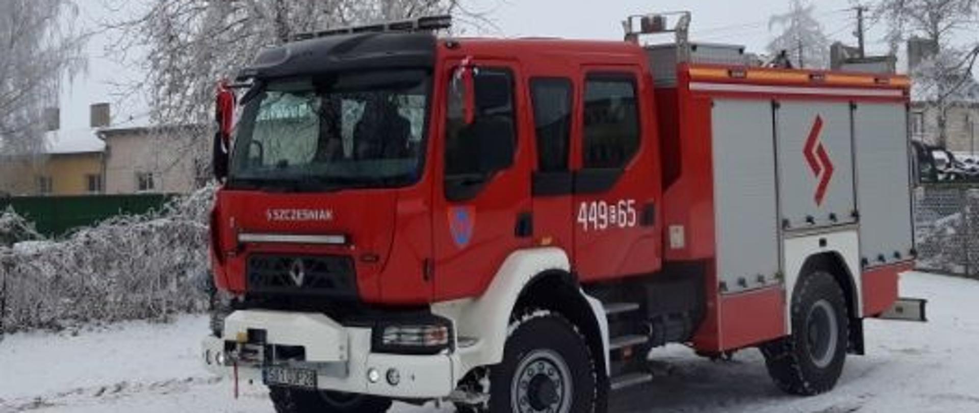 Na zdjęciu czerwony samochód strażacki. Zderzak i nadkole są w kolorze białym. Pozostałe dodatkowe elementy są czarne. Ustawiony jest w taki sposób, że widać przód i lewy bok. Samochód ma trzy srebrne skrytki z boku. Na drzwiach jest numer operacyjny 449E65. Na ziemi jest cienka warstwa śniegu. W tle widać budynki, krzewy i drzewa. Jest widno.