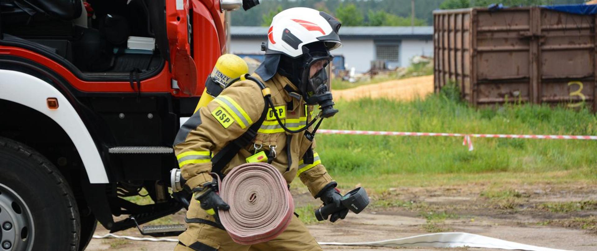 Strażak w pełnym umundurowaniu trzymający w prawym ręku wąż, w lewym kamerę termowizyjną 