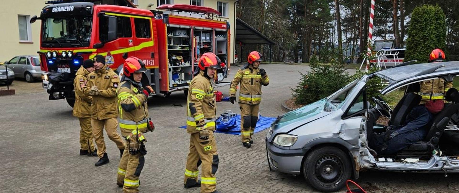 Grupa strażaków w mundurach bojowych i hełmach, przygotowują sprzęt przy w raku samochodu. W tle samochód strażacki.