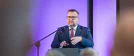 Na zdjęciu za mównicą stoi marszałek Szymon Ogłaza. W tle ścianka multimedialna. Na dole zdjęcia widać siedzących tyłem innych uczestników konferencji.