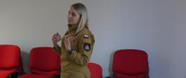 Świetlica komendy. Na tle ściany i ustawionych przy niej krzesłach stoi pani psycholog, która prowadzi psychoedukację. 