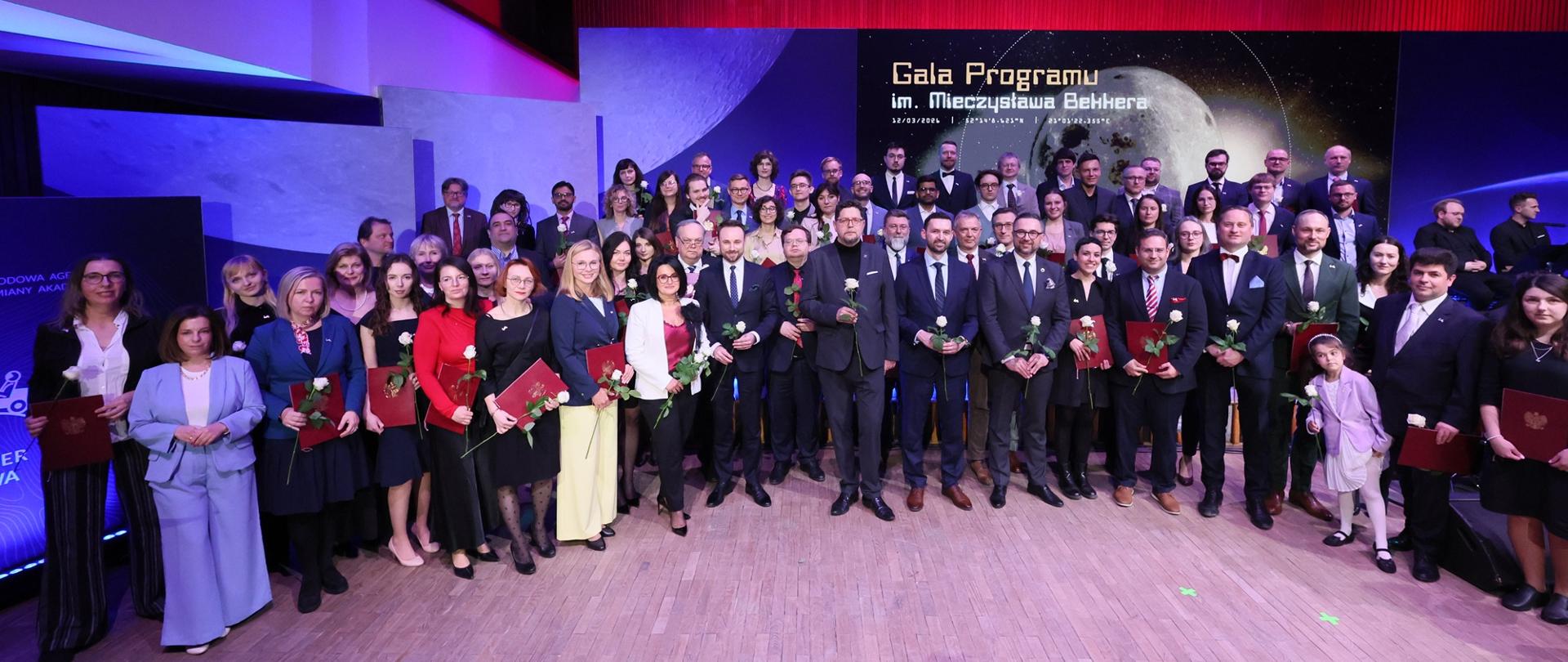Zdjęcie zbiorowe, na podwyższeniu z przodu dużej sali stoi kilkadziesiąt osób, pośrodku minister Kulasek, wiceminister Szeptycki i dwóch mężczyzn w garniturach, wszyscy trzymają w rękach białe kwiaty, za nimi wielki ekran z napisem Gala Programu im. Mieczysława Bekkera, w tle oświetlona na czerwono ściana.
