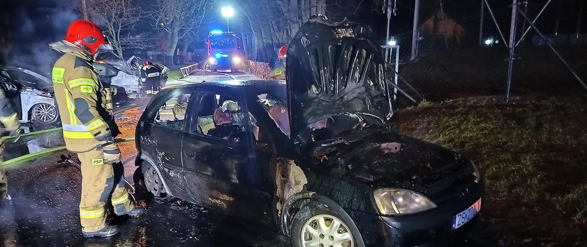 Strażak stoi przy spalonym samochodzie osobowym, z boku przody dwóch kolejnych objętych pożarem samochodów, w tle samochód ratowniczo-gaśniczy.