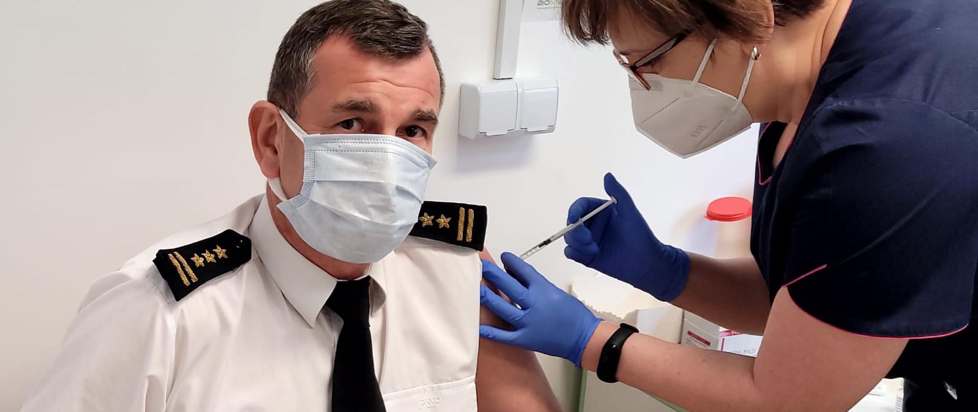 W lewej części zdjęcia st. bryg. mgr inż. Janisław Buller - Komendant Miejski Państwowej Straży Pożarnej w Bydgoszczy siedzący na krześle ubrany w białą koszulę z krótkim rękawem umundurowania wyjściowego PSP, czarny krawat z logo PSP, czarne pagony ze złotymi naszyciami stopnia służbowego starszego brygadiera, na twarzy maseczka. Obok Komendanta nachyla się stojąca pielęgniarka dokonująca szczepienia w lewy bark, pielęgniarka w ciemnym granatowym ubraniu, na twarzy maseczka. 