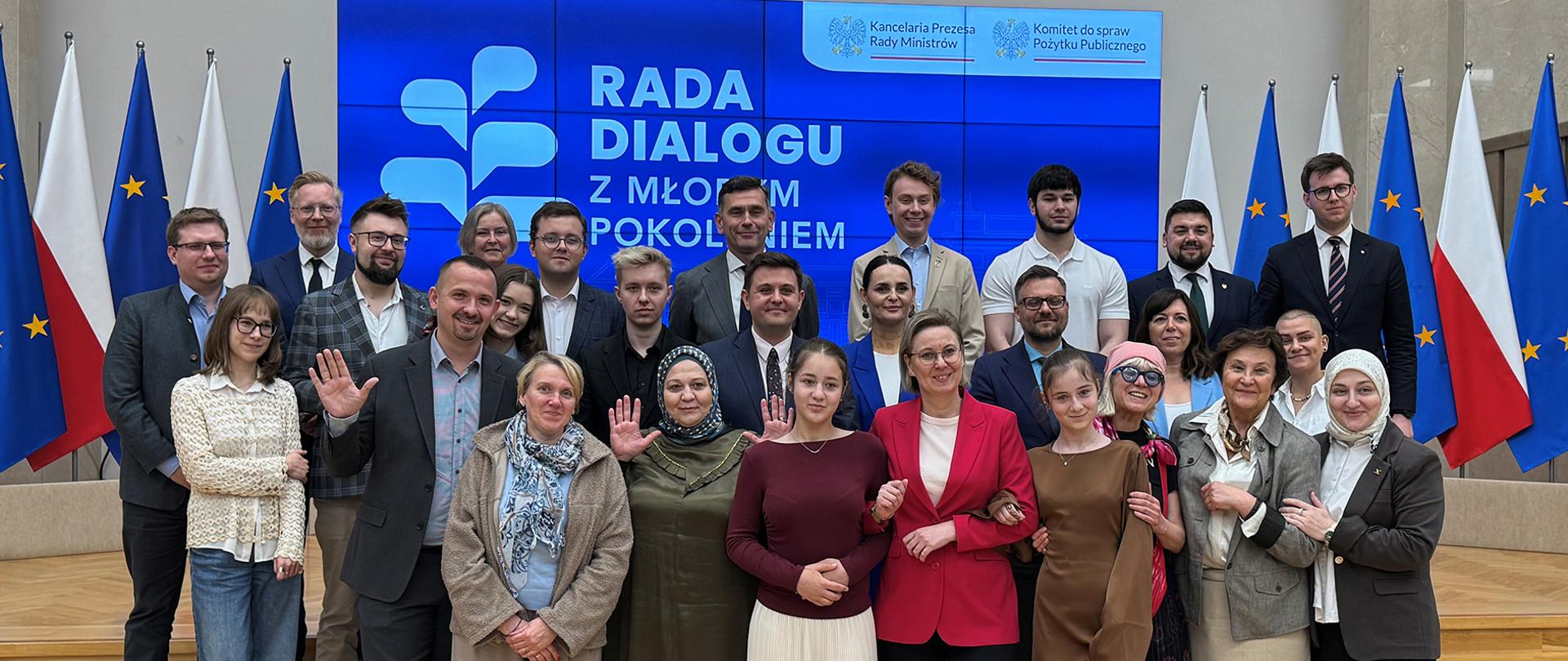 Zdjęcie grupowe. Uczestnicy wydarzenia pozują do wspólnego zdjęcia. W pierwszym rzędzie widoczna między innymi Sekretarz Stanu Adriana Porowska. W tle widoczny ekran, na którym wyświetlone jest logo Rady Dialogu z Młodym Pokoleniem. Po obu stronach ekranu stoją flagi Polski i Unii Europejskiej.