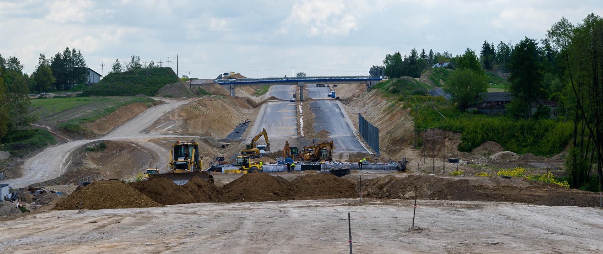 Budowa drogi ekspresowej w zaawansowanym etapie – widok na szeroki pas ziemi przygotowany pod nawierzchnię, z maszynami budowlanymi (koparki, spycharki) pracującymi w tle. Po obu stronach widoczne skarpy i ekrany akustyczne, a w oddali wiadukt nad przyszłą trasą. Otoczenie to tereny zielone z drzewami i zabudową w tle.