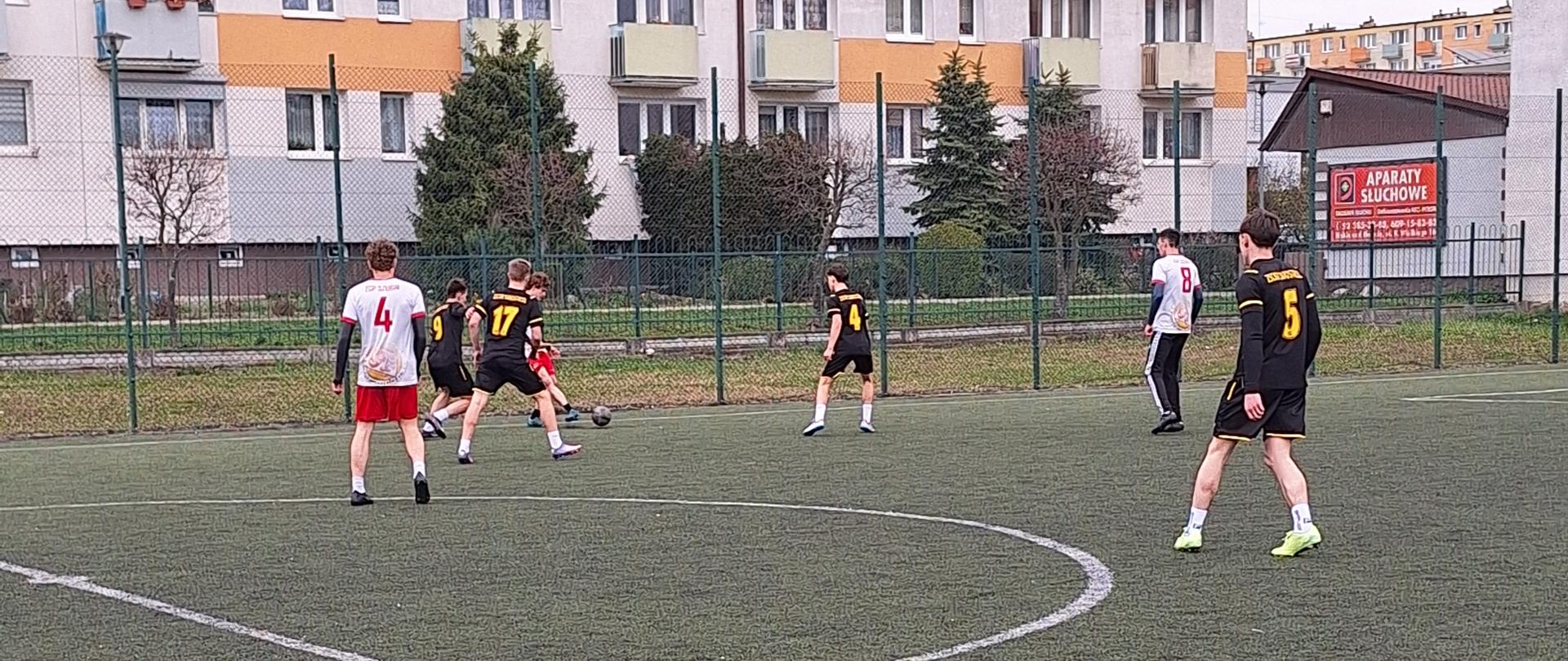 Młodzi piłkarze w czarnych i białych strojach sportowych podczas dynamicznej akcji na boisku typu Orlik. W tle widać wysoki, kolorowy blok mieszkalny oraz ogrodzenie boiska. Na pierwszym planie wyraźne linie wyznaczające środek pola gry.
