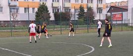 Szeroki kadr na boisko piłkarskie typu Orlik. Zawodnicy w czarnych strojach z żółtymi numerami (widoczny numer 17 oraz 5) otaczają przeciwnika w białej koszulce z numerem 4. Akcja toczy się na tle siatki ochronnej boiska i wielopiętrowego bloku mieszkalnego.