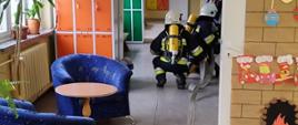 Zdjęcie przedstawia strażaków OSP podczas symulowanych działań na terenie szkoły. Po prawo naklejony na drzwi narysowany kominek z ogniem. po lewo stolik i dwa fotele a za nimi szafki szkolne.