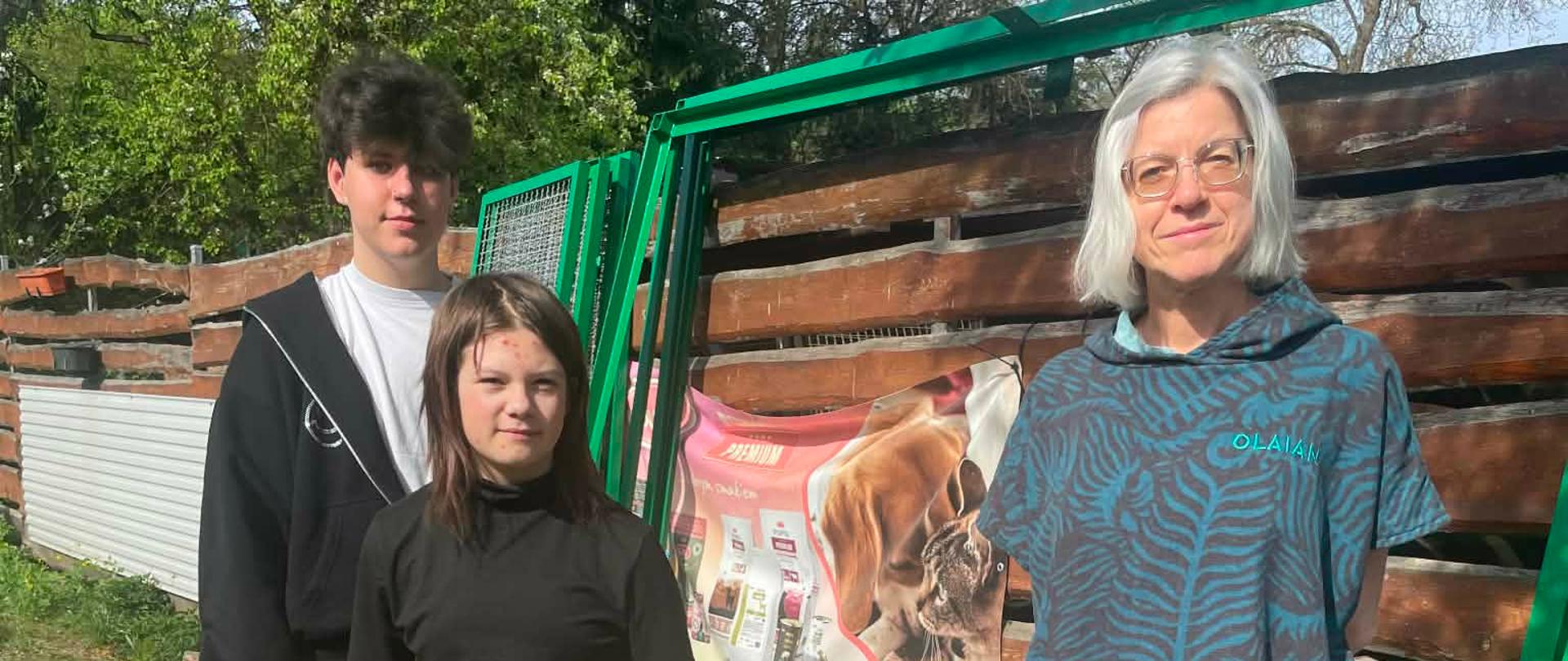 Na zdjęciu widać nauczycielkę i dwoje uczniów którzy stoją przed schroniskiem dla zwierząt a obok na palecie leżą dary dla tych zwierząt