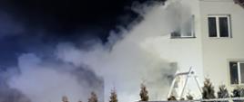 Zdjęcie przedstawia część budynku mieszkalnego dwurodzinnego objętego pożarem, gdzie z otworów okiennych wydobywa się dym