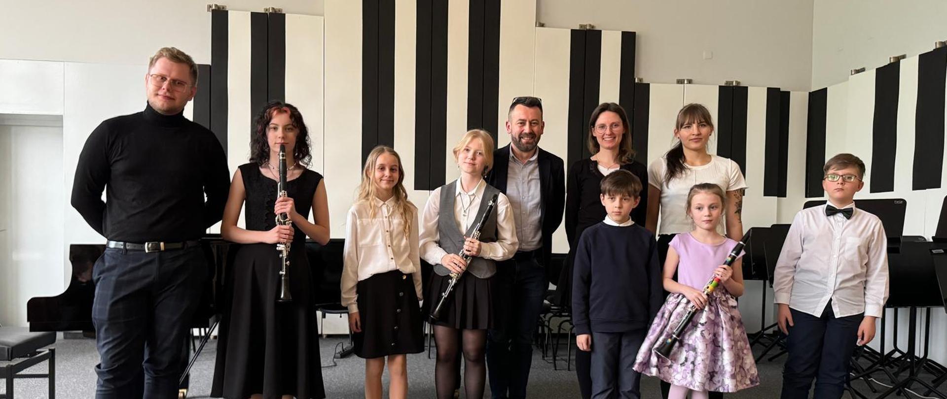 Siedmioro uczniów, kobieta i dwóch mężczyzn stoją na estradzie, troje trzyma klarnety.
