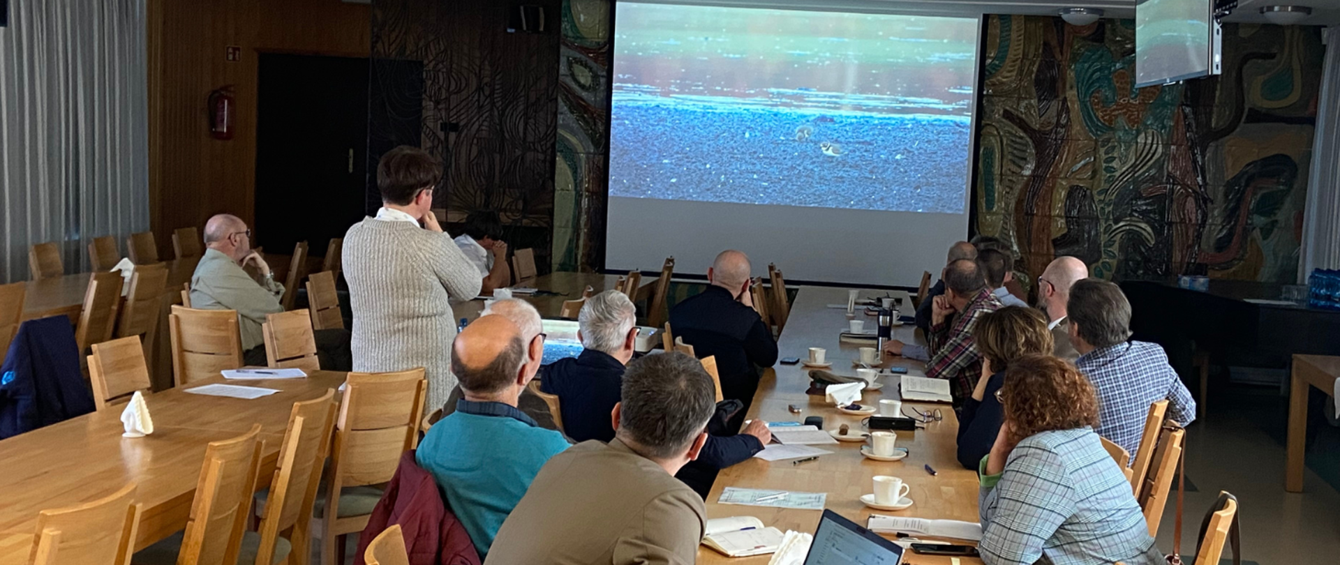 Dwa duże stoły, na których leżą dokumenty, wokół nich siedzą ludzie patrzą na wyświetloną prezentację. 