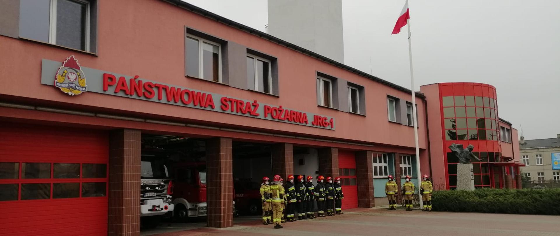 Na zdjęciu widzimy dwa szeregi strażaków ubranych w specjalne ubrania ochronne koloru piaskowego oraz koloru czarnego, na twarzach maski ochronne koloru białego. W środkowej części zdjęcia widzimy szereg ośmiu strażaków stojących pod budynkiem komendy. Bokiem zwrócony jest do nich dowódca wydający komendy. W prawej części zdjęcia widzimy drugi szereg złożony z trzech strażaków stojących pod masztem, zwróconych twarzą do dowódcy. Na głowach czerwone hełmy z cyfrą "1". Za szeregiem strażaków uwidoczniony jest maszt koloru białego uwieńczony biało-czerwoną flagą. Dalej w kadrze widzimy część budynku komendy wraz z pomnikiem strażaka trzymającego dziecko oraz roślinność dekoracyjną. Budynek komendy koloru łososiowego wraz z oszklonym wejściem głównym koloru czerwonego. Z lewej strony kadru widzimy pięć alarmowych bram wyjazdowych koloru czerwonego, z czego trzy bramy są otwarte. W otwartych bramach widać fragmenty pojazdów pożarniczych. Powyżej bram uwidoczniony jest napis koloru czerwonego "Państwowa Straż Pożarna JRG-1 oraz logo Państwowej Straży Pożarnej. Z prawej strony kadru w oddali widzimy budynek mieszkalny ( kamienica dwukondygnacyjna ) koloru szarego oraz cześć banera reklamowego. 