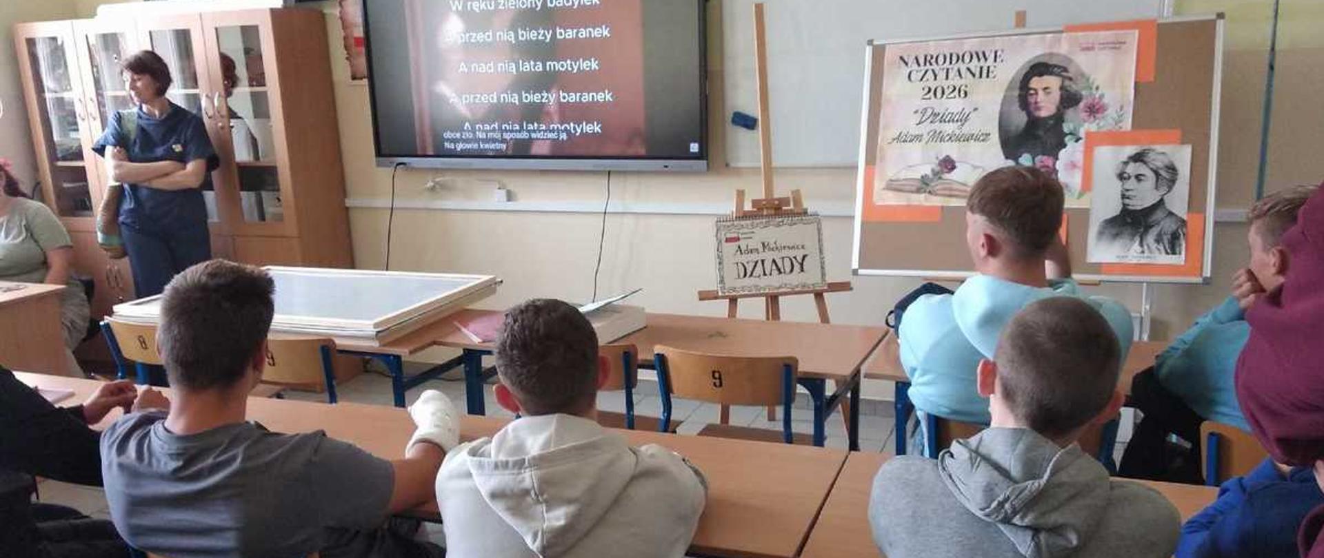 Zdjęcie przedstawia salę lekcyjną w polskiej szkole, w której odbywa się wydarzenie z cyklu Narodowe Czytanie 2026. W ławkach tyłem do obiektywu siedzą uczniowie, a ich uwaga skierowana jest na przód klasy.Pierwszy plan (Uczniowie w ławkach)W dolnej części kadru widoczne są plecy i tyły głów kilkorga nastolatków siedzących przy szkolnych ławkach na drewnianych krzesłach z wyraźnie namalowaną czarną cyfrą „9”.Po lewej stronie chłopak w ciemnoszarej koszulce opiera się o blat.Obok niego, w środkowej części, siedzi uczeń w jasnoszarej bluzie z kapturem, z założonymi na plecach rękami.Po prawej stronie widać kolejnego chłopca w szarej bluzie z kapturem oraz fragmenty sylwetek innych uczniów.Drugi plan (Sceneria i dekoracje)Na środku sali, tuż przed tablicami, stoi mała drewniana sztaluga, na której oparto białą tabliczkę z napisem wykonanym ozdobną czcionką: „Adam Mickiewicz DZIADY”.Po prawej stronie znajduje się duży stojak z tablicą informacyjną wydarzenia. Widnieje na niej nagłówek: „NARODOWE CZYTANIE 2026 »Dziady« Adam Mickiewicz” wraz z portretem poety oraz ilustracją przedstawiającą otwartą książkę i postać kobiety w ciemnej sukni na tle różowych kwiatów. Obok, na tej samej tablicy, przypięty jest mniejszy, czarno-biały portret Adama Mickiewicza.Tło (Wyposażenie sali)W głębi klasy, na ścianie, wiszą dwa duże ekrany:Po lewej stronie znajduje się włączony ekran multimedialny. Wyświetla on slajd z fragmentem tekstu (wiersza) na ciemnoczerwonym tle. Widoczne są m.in. słowa: „Na głowie kwietny ma wianek / W ręku zielony badylek / [...] A nad nią lata motylek”.Po prawej stronie wisi tradyzyjna biała tablica suchościeralna z zapisanymi w rogu kredą cyframi lub wzorami. Nad nią wisi tradycyjny drewniany krzyż.W tle po lewej stronie, przy brązowej szafie bibliotecznej, stoi tyłem do okna kobieta (prawdopodobnie nauczycielka) w ciemnoniebieskim stroju, z założonymi rękami, która obserwuje klasę. Pomieszczenie jest jasne, oświetlone światłem dziennym wpadającym przez duże okna po lewej stronie.