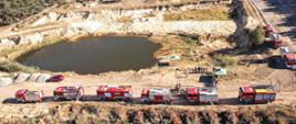Uczestnicy ćwiczeń w trakcie odprawy. Po boku znajdują się pojazdy pożarnicze. Zdjęcie wykonane na terenie żwirowni.
