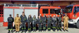 Kursanci i kursantki wraz z egzaminatorami pozują do zdjęcia grupowego na t;e samochodów pożarniczych. 