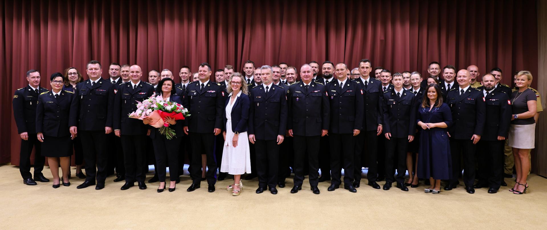 Na zdjęciu znajduje się duża grupa uczestników spotkania – funkcjonariuszy Państwowej Straży Pożarnej oraz pracowników Komendy Wojewódzkiej PSP w Warszawie. Osoby stoją w kilku rzędach przed bordowymi kotarami; większość funkcjonariuszy ma na sobie ciemne mundury służbowe, a część uczestników ubrana jest po cywilnemu. W centrum grupy widoczna jest kobieta z bukietem kwiatów, a w pierwszym rzędzie stoją także osoby uczestniczące w uroczystości. Zdjęcie ma oficjalny, pamiątkowy charakter i dokumentuje całe spotkanie pożegnalne.