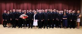Na zdjęciu znajduje się duża grupa uczestników spotkania – funkcjonariuszy Państwowej Straży Pożarnej oraz pracowników Komendy Wojewódzkiej PSP w Warszawie. Osoby stoją w kilku rzędach przed bordowymi kotarami; większość funkcjonariuszy ma na sobie ciemne mundury służbowe, a część uczestników ubrana jest po cywilnemu. W centrum grupy widoczna jest kobieta z bukietem kwiatów, a w pierwszym rzędzie stoją także osoby uczestniczące w uroczystości. Zdjęcie ma oficjalny, pamiątkowy charakter i dokumentuje całe spotkanie pożegnalne.