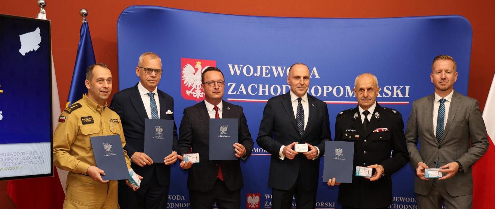 Zdjęcie z uroczystości w Urzędzie Wojewódzkim podczas inauguracji "czujne Zachodniopomorskie. Pięć osób w tym dwie w mundurach strażackich, jeden w służbowym koloru pisakowego drugi w mundurze wyjściowym ochotniczej straży pożarnej koloru granatowego. pozostali w garniturach. Wszyscy stoją na tle niebieskiej ścianki z napisem wojewoda zachodniopomorski z Godłem Polski, prezentując czujki tlenku węgla oraz trzymając granatowe teczki z podpisanym porozumieniem w sprawie realizacji akcji „Czujne Zachodniopomorskie”.