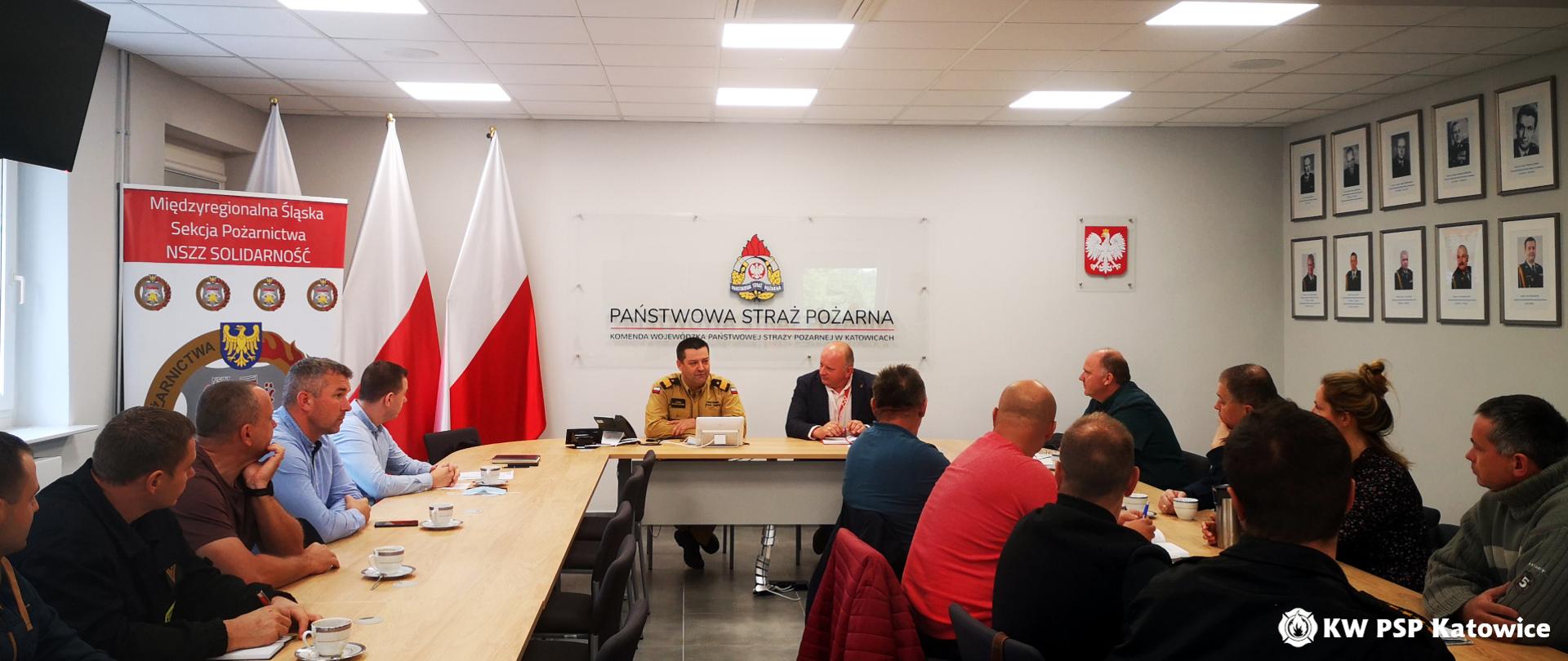 Przy stole prezydialnym w sali sztabowej Komendy Wojewódzkiej PSP w Katowicach od lewej strony siedzi Śląski Komendant Wojewódzki PSP - nadbryg. Jacek Kleszczewski oraz Pan Roman Marekwica Wiceprzewodniczący Międzyregionalnej Śląskiej Sekcji Pożarnictwa NSZZ „Solidarność”. Po dwóch stronach członkowie związku. W tle napis Państwowa Straż Pożarna, Komenda Wojewódzka PSP, po lewej trzy biało-czerwone flagi i baner NSZZ Solidarność.