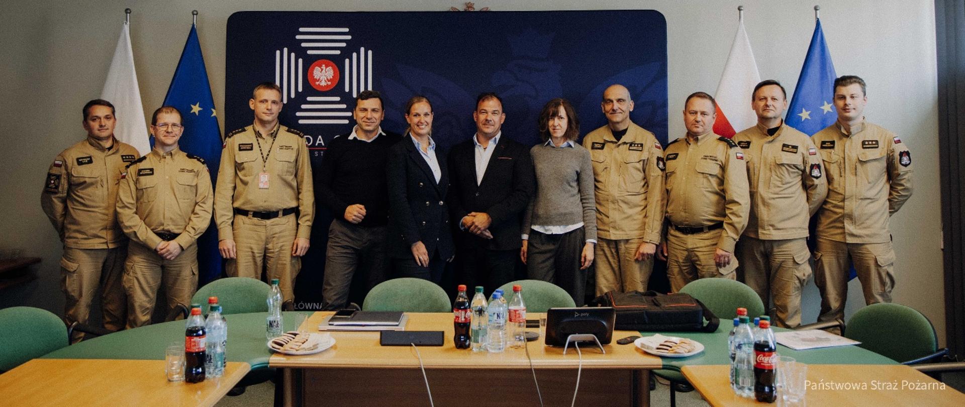 Grupa strażaków PSP w żółtych mundurach służbowych oraz osób cywilnych pozuje wspólnie do zdjęcia grupowego w sali konferencyjnej Komendy Głównej PSP. W tle widoczne są flagi Polski i Unii Europejskiej oraz logo PSP.