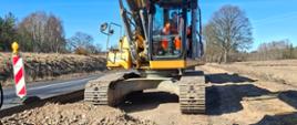 Na zdjęciu znajduje się duża koparka gąsienicowa ustawiona na rozkopanym odcinku drogi. Wokół maszyny leżą kopce ziemi i kamieni, a przednia łyżka jest częściowo widoczna na pierwszym planie. Po lewej stronie widać znak tymczasowej organizacji ruchu, a w tle rozciąga się teren z niską roślinnością i drzewami pod bezchmurnym niebem.