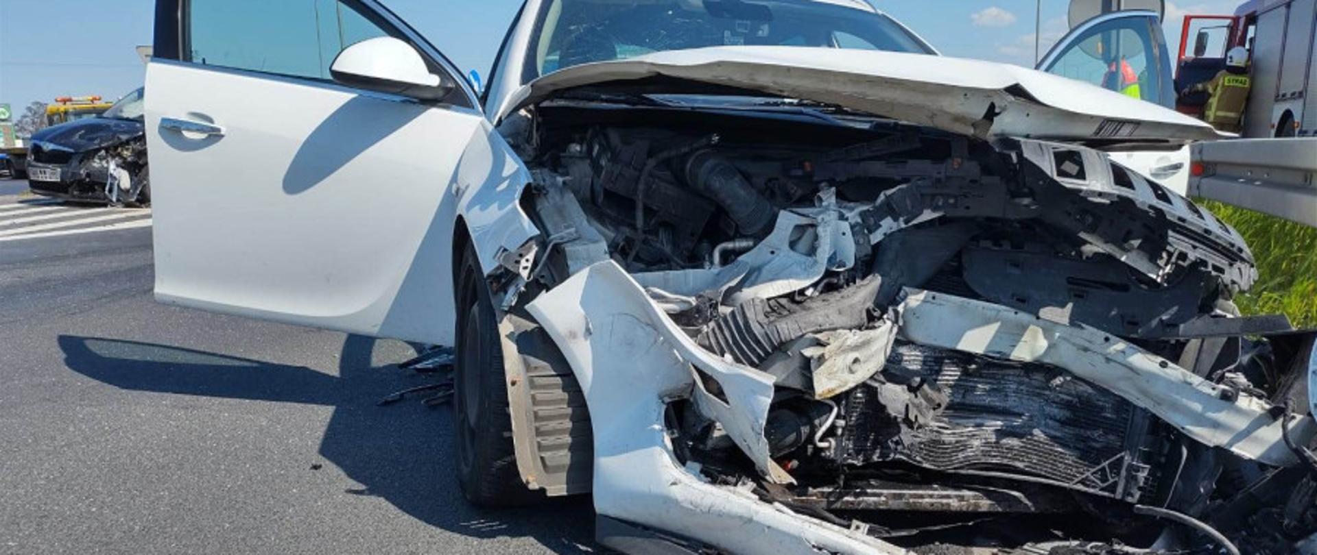 Zdjęcie przedstawia samochód osobowy koloru białego po zderzeniu. Zniszczony przód pojazdu