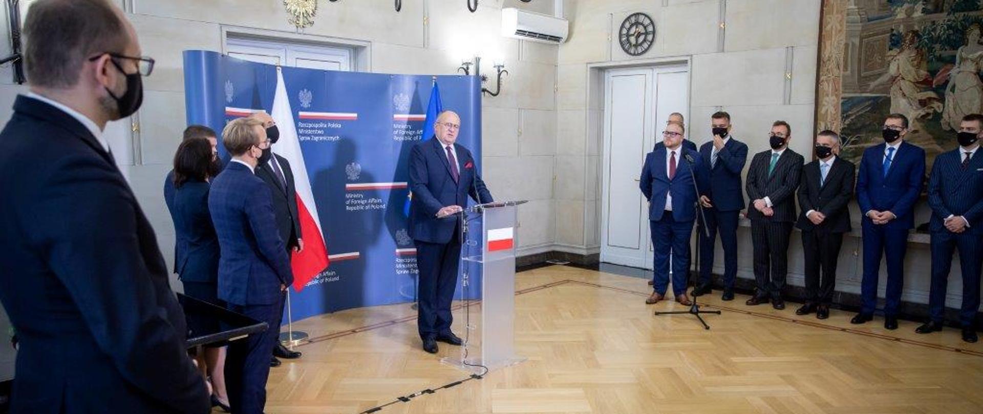 Minister Rau stoi przed mikrofonem w towarzystwie grupy dyplomatów. Za nim ścianka z logiem MSZ.