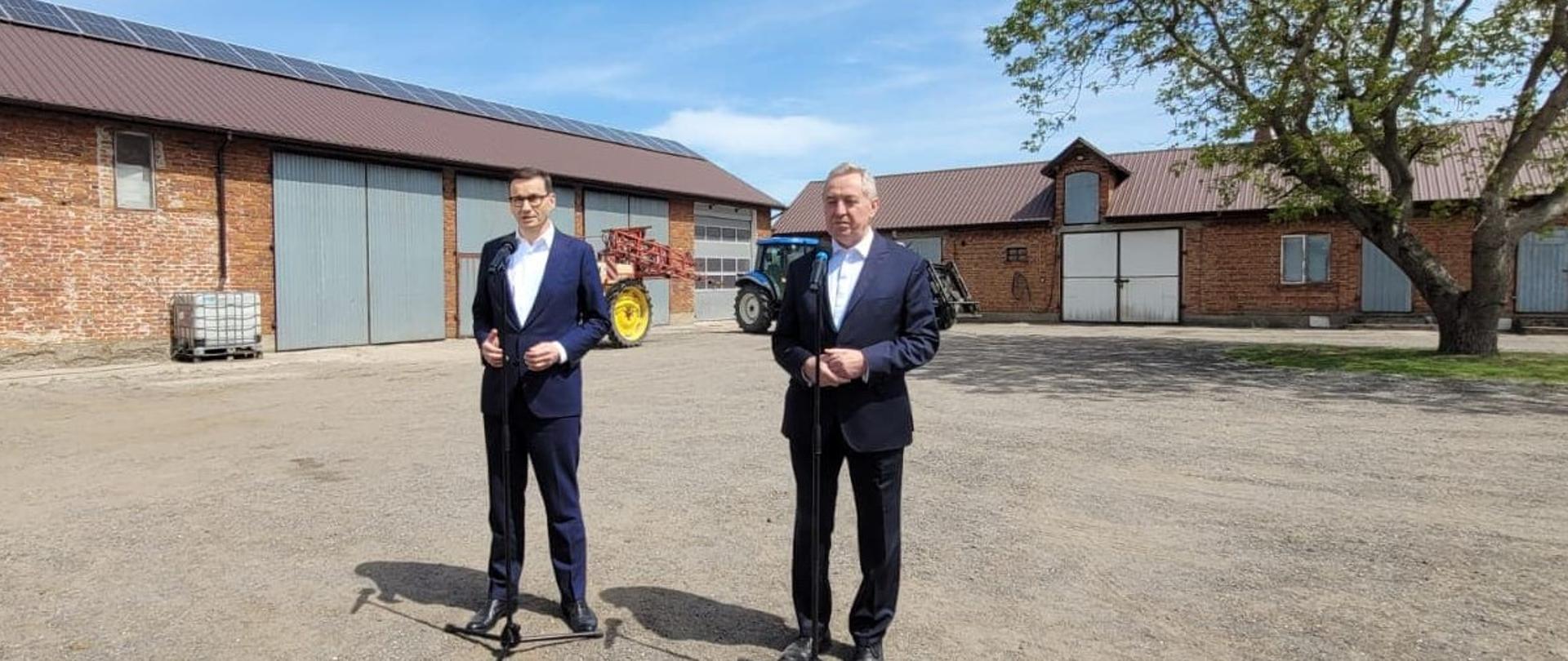 premier Mateusz Morawiecki oraz wicepremier, minister rolnictwa i rozwoju wsi Henryk Kowalczyk stojący na podwórku gospodarstwa rolnego