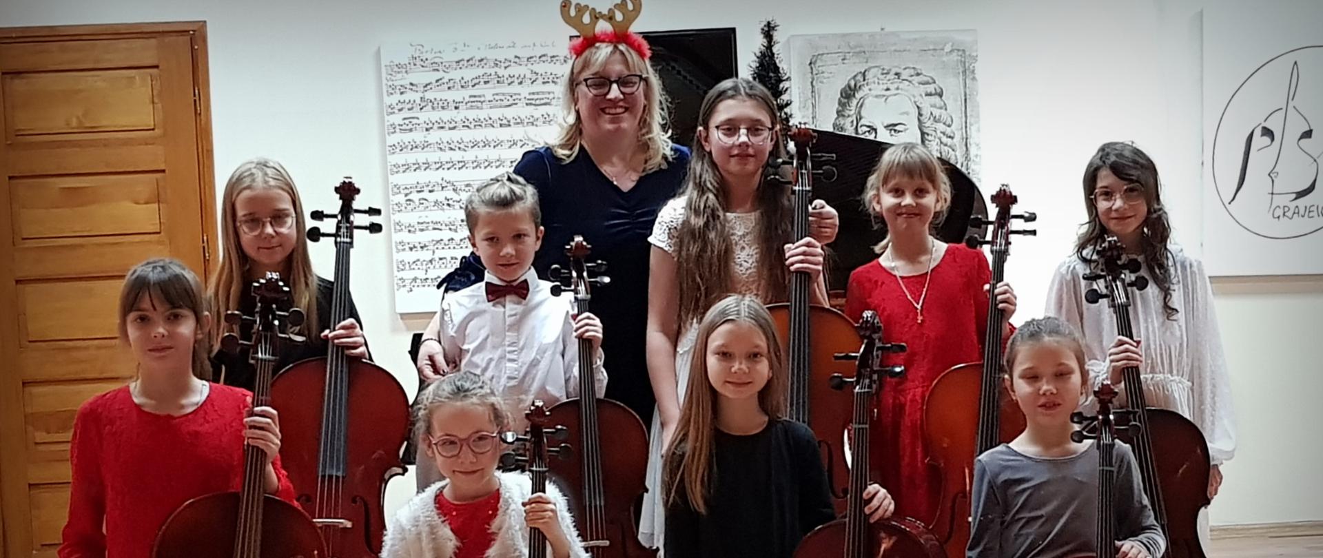 Uczniowie stoją, każdy z wiolonczelą, za nimi nauczycielka, w tle widać obrazy