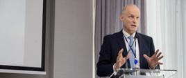 Zdjęcie przedstawia mężczyznę wiceministra Wojciecha Zajączkowskiego w garniturze stojącego na mównicy z polską flagą