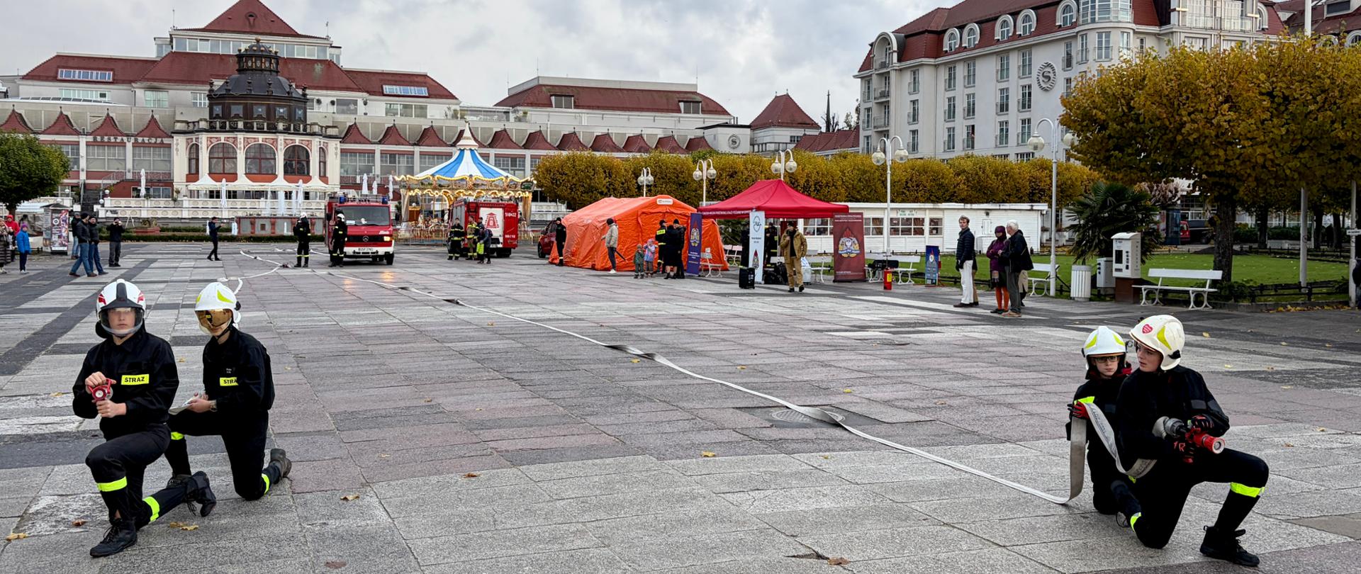 Strażacy w hełmach i umundurowaniu ćwiczebnym rozwijają węże pożarnicze na placu przed Domem Zdrojowym w Sopocie. W tle widać czerwone i pomarańczowe namioty PSP, wozy strażackie oraz budynki z czerwonymi dachami. Kilka osób obserwuje pokaz z chodnika.