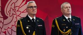 Zdjęcie przedstawia dwóch wysokich rangą oficerów Państwowej Straży Pożarnej w pełnych mundurach wyjściowych. Mężczyźni stoją na baczność obok siebie na czerwonym dywanie, na tle oficjalnej ścianki reprezentacyjnej. Kadrowanie obejmuje sylwetki od stóp w górę. Oficer po lewej stronie st. bryg. Piotr Grzyb stoi wyprostowany, patrząc lekko w górę i w prawą stronę. Ma krótkie, szpakowate włosy oraz okulary korekcyjne w ciemnych oprawkach. Nosi ciemnogranatowy mundurowy garnitur garnizonowy. Na prawym ramieniu ma przypięty złoty sznur galowy. Na naramiennikach (pagonach) widoczne są złote dystynkcje oficerskie starszego brygadiera (trzy gwiazdki i dwie belki). Na lewej piersi przypięty jest rząd wielokolorowych baretek odznaczeń, a poniżej klapy po prawej stronie znajduje się odznaka pamiątkowa. Nosi czarne, eleganckie półbuty. Oficer po prawej stronie to Komendant Powiatowy PSP w Wołowie stoi na baczność, patrząc na wprost przed siebie. Ma krótkie, jasne włosy i regularne rysy twarzy. Nosi identyczny krój ciemnogranatowego munduru ze złotym sznurem galowym na prawym ramieniu. Na pagonach widoczne są cztery złote gwiazdki. Na lewej piersi ma pojedynczą baretkę, a na prawej kieszeni przypiętą odznakę Szkoły Aspirantów PSP oraz inne odznaczenia. Na nogach ma czarne, skórzane półbuty. W lewym dolnym rogu kadru widoczny jest fragment drewnianej mównicy. Na jej froncie umieszczono duże, kolorowe logo Państwowej Straży Pożarnej (orzeł na tle płomieni, otoczony napisem „Straż Pożarna”). Ścianka w tle: Wielkoformatowa, czerwona ścianka z geometrycznym, bordowo-czerwonym wzorem. Oficerowie stoją na jaskrawoczerwonym dywanie reprezentacyjnym.