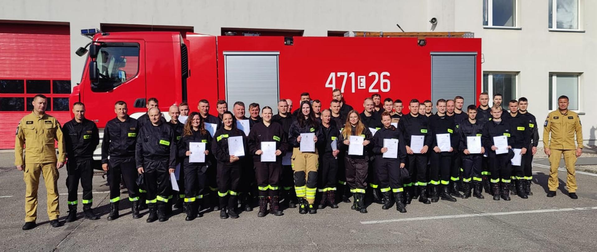 Grupa strażaków w ubraniach koszarowych koloru czarnego trzymająca zaświadczenia ukończenia kursu. Dwóch funkcjonariuszy w ubraniach służbowych koloru piaskowego na tle samochodu bojowego i budynku komendy powiatowej