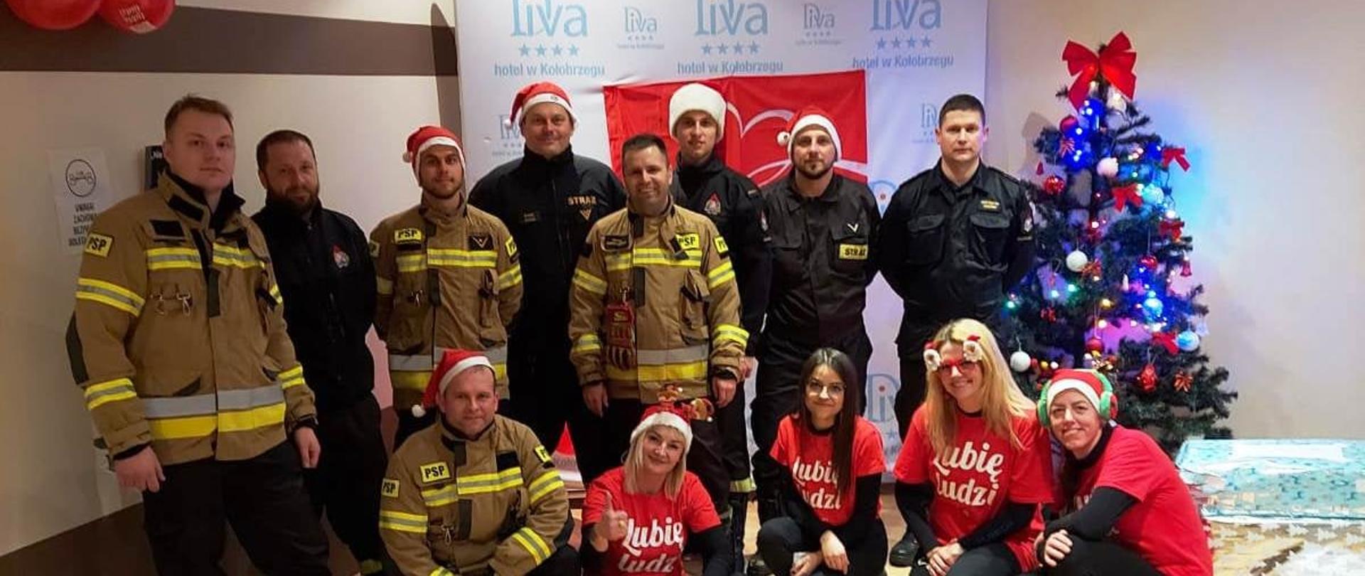 Strażacy i wolontariusze ubrani w czerwone stroje i czapki na tle baneru z logo "Szlachetnej Paczki". Za ich plecami choinka świąteczna. Obok przygotowane paczki.