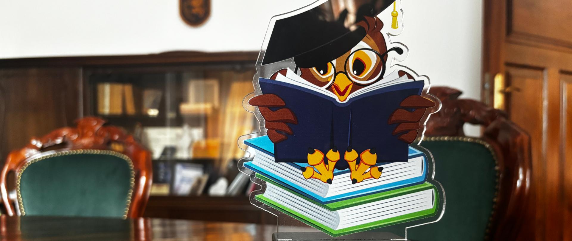 Nagroda w kształcie sowy w okrągłych okularach i uniwersyteckiej czapce siedząca na zielonej i niebieskiej książce, trzymająca w skrzydłach otwartą, granatową książkę. Pod nią złota tabliczka z napisem "Przyjaciel Zespołu Szkół w Lotyniu". Nagroda umiejscowiona na brązowym blacie. W tle dwa krzesła z zielonym obiciem, drzwi oraz regał z książkami.