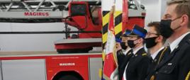 na zdjęciu sześciu strażaków w mundurach galowych stojących bokiem do robiącego zdjęcie, strażacy stoją na uroczystej zbiórce w garażu, trzech z nich stoi w poczcie sztandarowym, w tle widać bok samochodu z drabiną mechaniczną