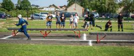 Na zdjęciu dwóch strażaków w strojach sportowych biegnących po bieżni i równoważni, w tle kibice obserwujący bieg zza taśmy ostrzegawczej i zaparkowane samochody.