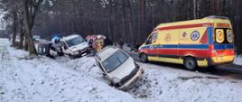 Dwa samochody osobowe oraz jeden dostawczy znajdują się w rowie. Żółta karetka pogotowia ratunkowego stoi i na jezdni. W tle las, jeden samochód strażacki, inne pojazdy oraz strażacy. Pora zimowa, śnieg na powierzchni.