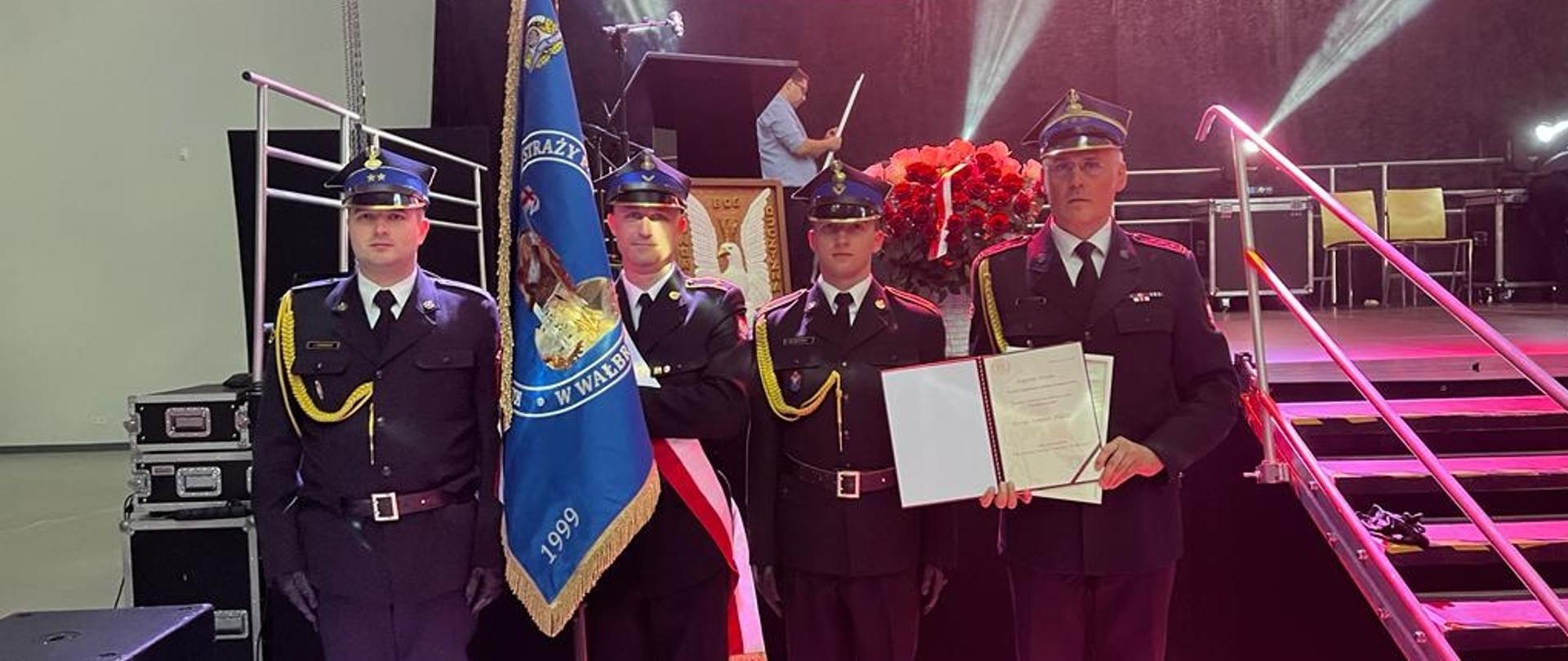 Czterech strażaków w mundurach wyjściowych stoi na baczność, jeden z nich trzyma niebieski sztandar. Z prawej strony Komendant Miejski PSP w Wałbrzychu trzyma w ręku odznaczenia. W tle biało czerwona flaga