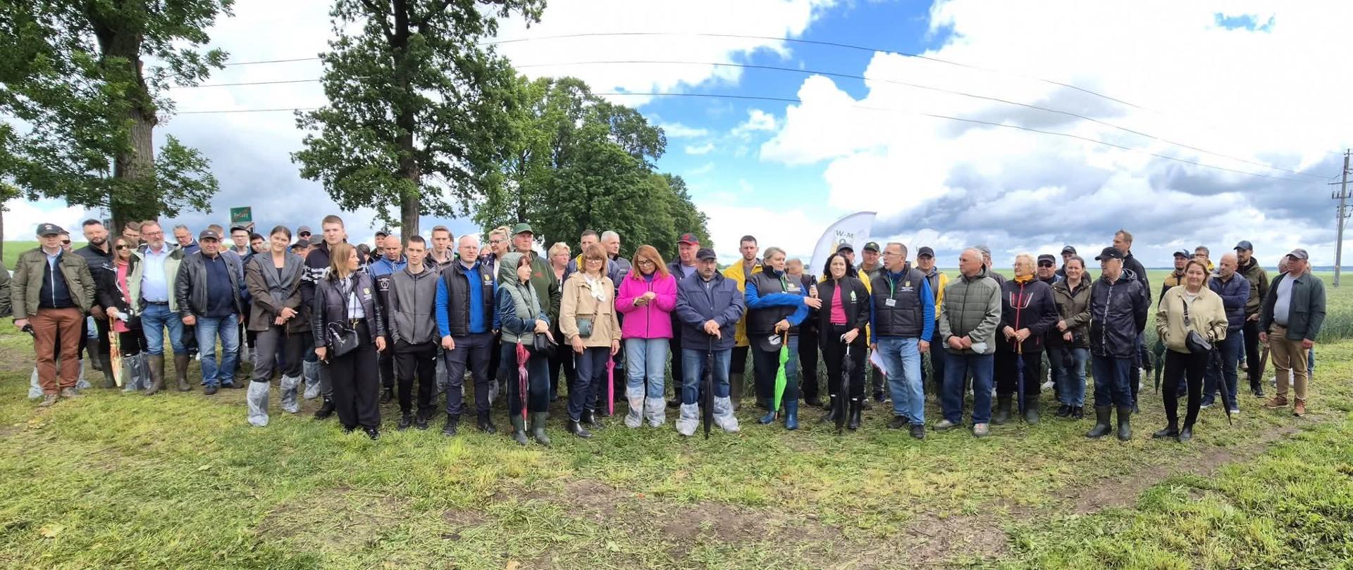 Duża grupa osób pozuje do zdjęcia na tle pól uprawnych częściowo zachmurzonego nieba