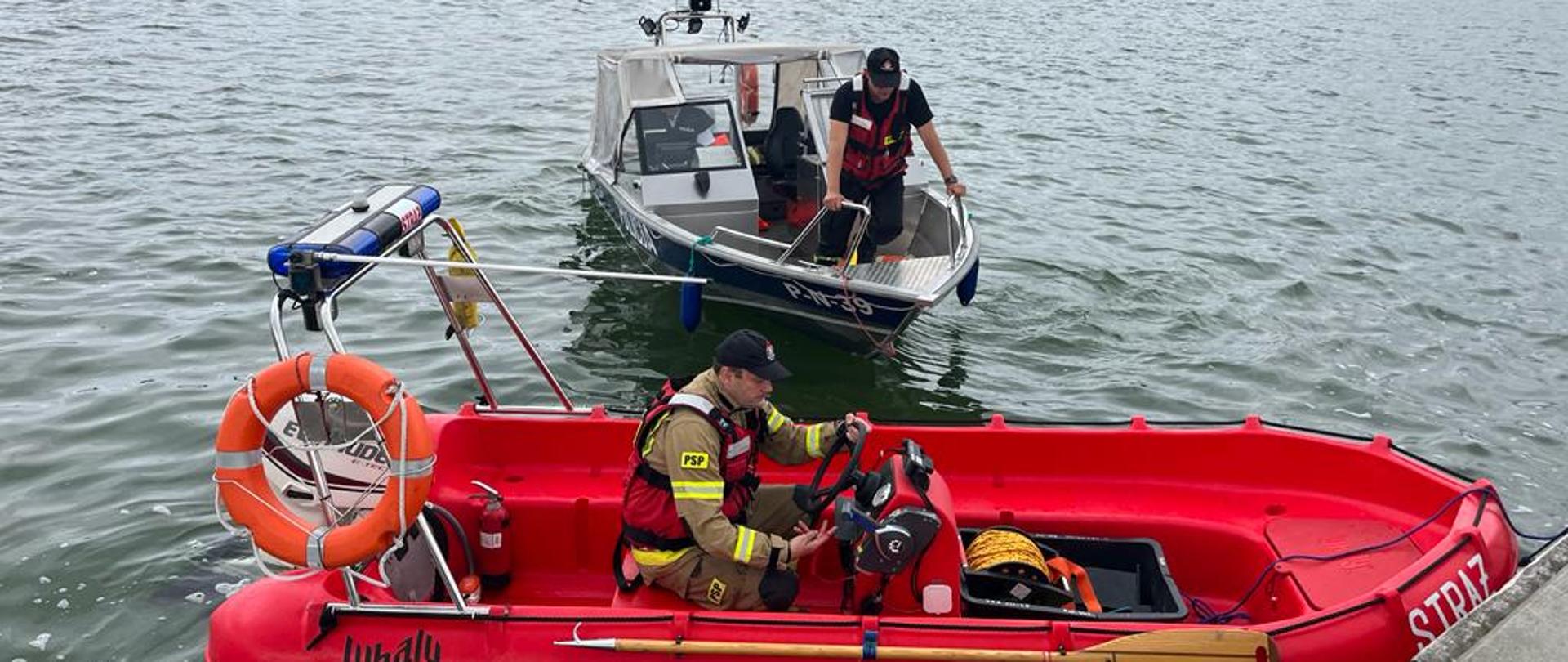 Dwie łodzie jedna w kolorze czerwonym z napisem STRAŻ i w oddali z napisem POLICJA zacumowane przy brzegu pomostu. Na łodziach widoczne osoby , strażak i policjant.
