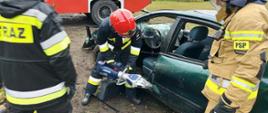 Strażak przy użyciu rozpieracza próbuje zerwać zawiasy we wraku samochodu. Z boku stoją strażacy i obserwują ćwiczenie