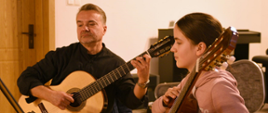Zdjęcie przedstawia od lewej strony mężczyznę trzymającego gitarę oraz uczennicę obserwującą zapis nutowy na pulpicie do nut na tle drzwi do sali i beżowej ściany.
