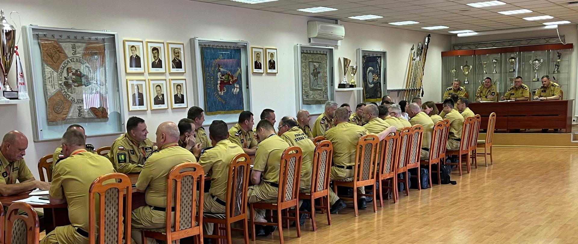 strażacy w mundurach w kolorze piaskowym oraz kobieta w białej bluzce siedzą przy stole w sali, za nimi ścianie na której wiszą gabloty ze sztandarami, zdjęcia oraz stoją puchary na półkach