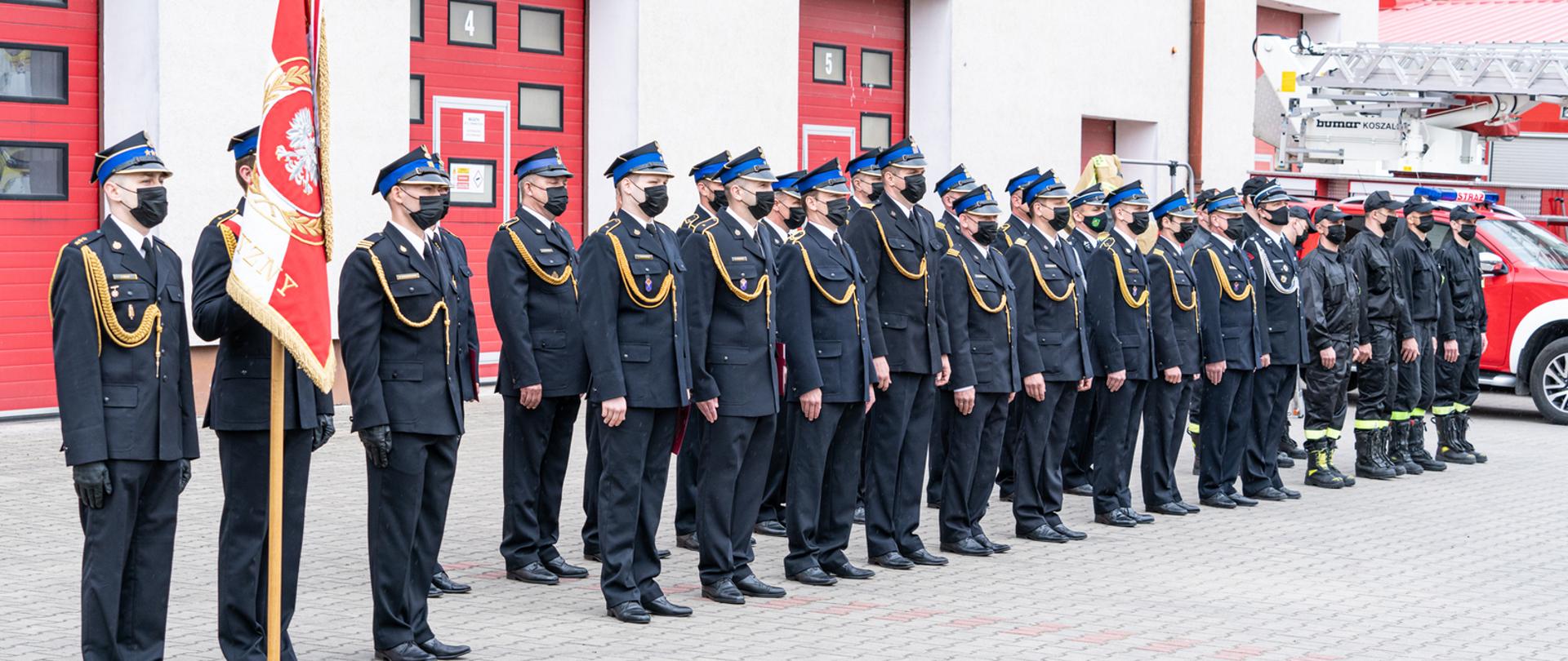 Zdjęcie zrobione na placu komendy przed budynkiem strażackim. W dwuszeregu ustawieni są strażacy w czarnym mundurze. Na czele stoi poczet sztandarowy ze sztandarem KP PSP w Kętrzynie.