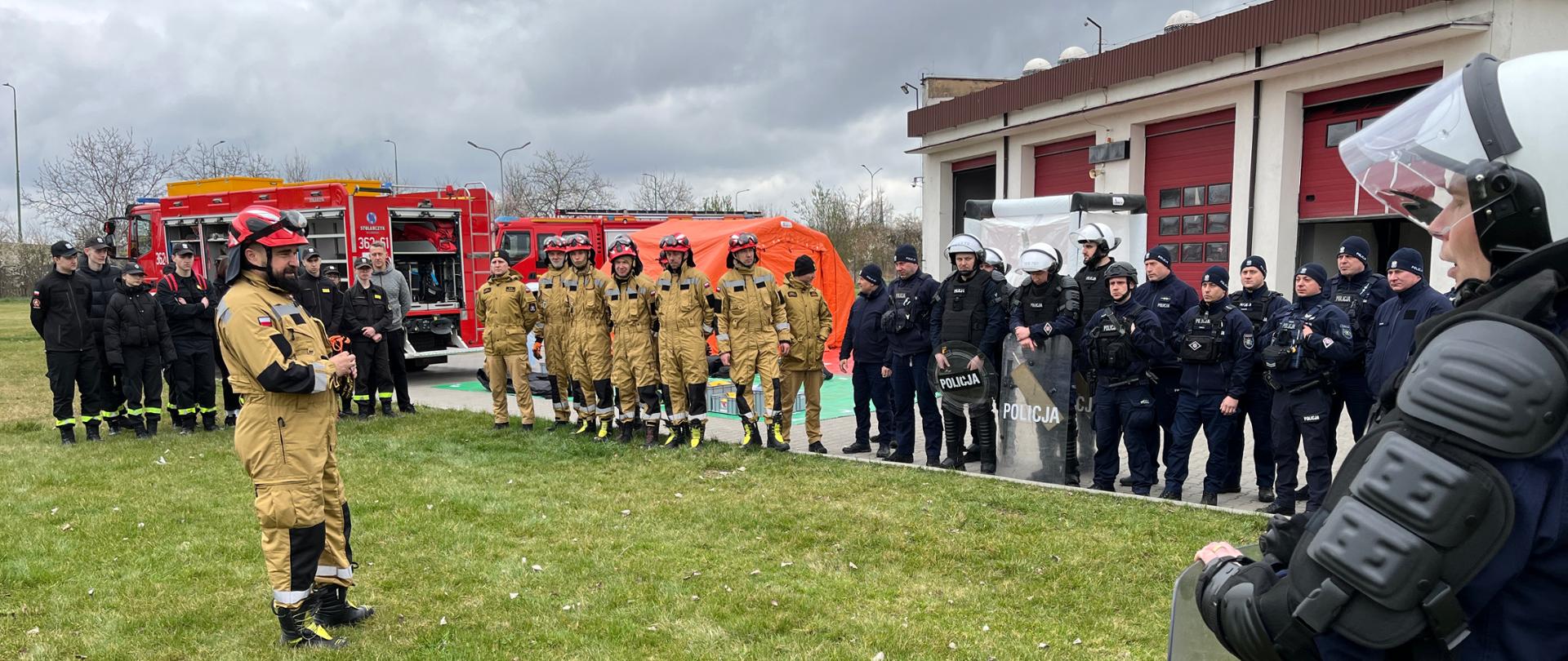 Zdjęcie przedstawia wspólne ćwiczenia służb mundurowych odbywające się na terenie jednostki straży pożarnej. Na fotografii można wyróżnić kilka grup uczestników i elementów wyposażenia: Strażacy: Widoczna jest grupa strażaków w piaskowych ubraniach specjalnych i hełmach, stojąca w szeregu, oraz drugi zespół w czarnych mundurach koszarowych. Jeden ze strażaków stoi przed grupą, prawdopodobnie instruując uczestników. Policjanci: Po prawej stronie zdjęcia znajduje się zwarta grupa funkcjonariuszy policji w ciemnogranatowych mundurach, wyposażonych w kamizelki taktyczne, kaski z przyłbicami oraz tarcze ochronne. Pojazdy i sprzęt: W tle zaparkowane są czerwone wozy strażackie z otwartymi skrytkami sprzętowymi oraz rozstawiony pomarańczowy namiot pneumatyczny. Otoczenie: Działania odbywają się na placu przed budynkiem garażowym z czerwonymi bramami wjazdowymi, przy pochmurnej pogodzie.