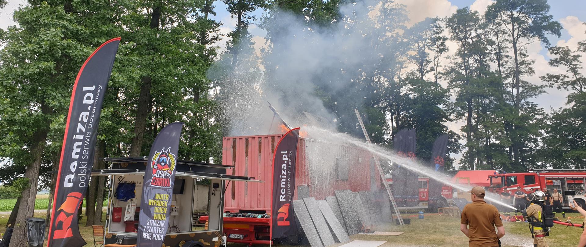 Zdjęcie w ciągu dnia wykonane podczas warsztatów ogniowych w Czartkach. Słonecznie. Na zdjęciu widać uczestników warsztatów. Podłoże trawiaste. 
