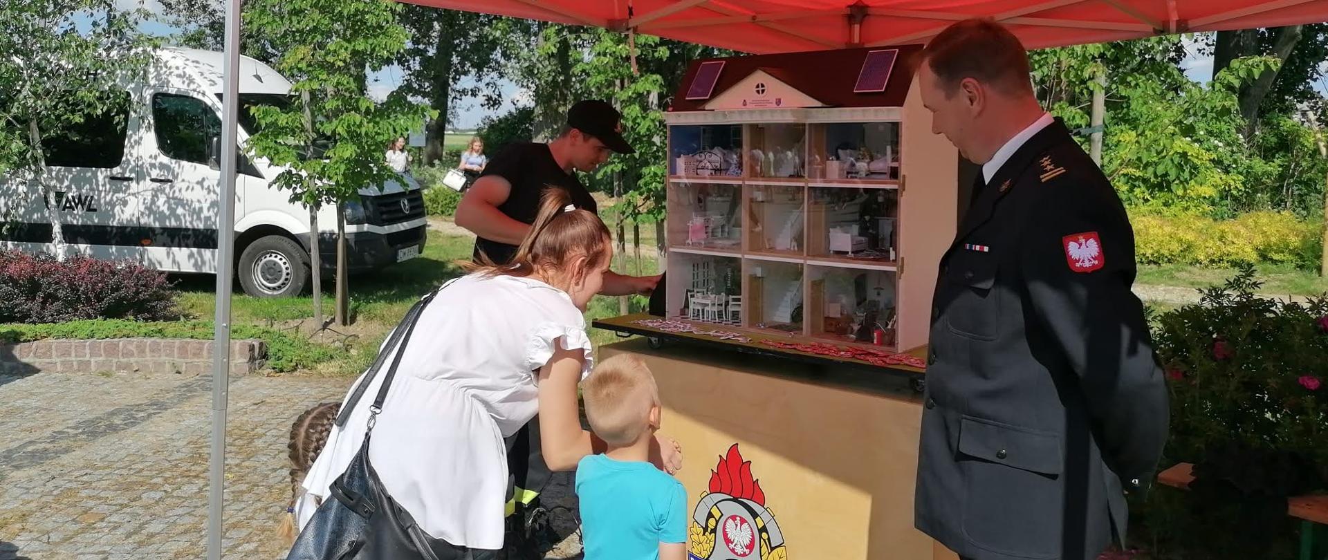 Strazak prezentuje mobilny symulator zagrożeń w domu jednorodzinnym. Całości przygląda się Komendant Powiatowy uczestniczący w pikniku. Pokaz obserwują dzieci z opiekunami.